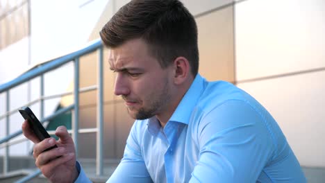 Profile-of-desperate-young-businessman-talking-on-smart-phone-and-sitting-on-stairs-near-office-building.-Close-up-of-upset-sad-manager-got-a-very-bad-news-on-cell-phone-and-crying-outdoor.-Side-view