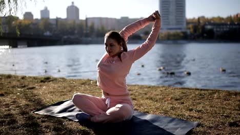 Mädchen-in-einen-Trainingsanzug-an-den-Ufern-eines-städtischen-Flusses-macht-Yoga-auf-einer-speziellen-Matte-und-hört-Musik.