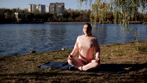 Mädchen-in-einen-Trainingsanzug-an-den-Ufern-eines-städtischen-Flusses-macht-Yoga-auf-einer-speziellen-Matte-und-hört-Musik.