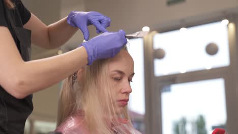 A-close-up-side-view-of-a-hairdresser-applying-dye-on-the-blonde's-hair.-The-camera-moves-slowly-top-down-and-stops-at-her-hands.-The-blonde-holds-a-phone,-looks-at-the-screen-and-types-a-message.