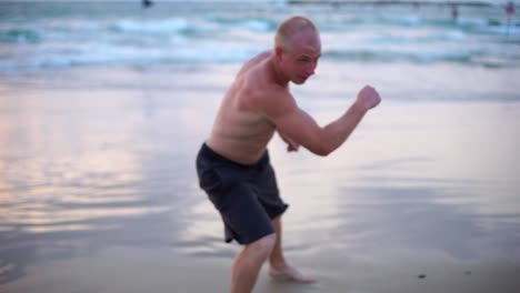 Joven-musculoso-hombre-practicando-ejercicios-de-boxeo-en-la-playa-del-mar.-Deportista-masculino-es-practica-defensa-personal-sola-cerca-de-la-orilla-del-mar.-Entrenamiento-de-artes-marciales-al-aire-libre-al-atardecer.-Lenta-de-cerca