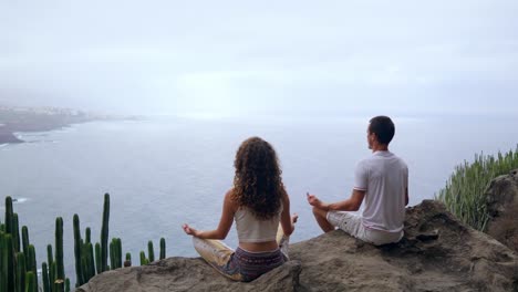 Un-hombre-y-una-mujer-sentada-en-la-cima-de-una-montaña-mirando-el-mar-sentado-en-una-piedra-meditando-en-posición-de-loto.-La-vista-desde-la-parte-posterior.-Islas-Canarias