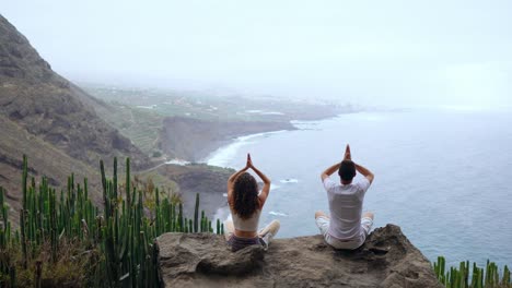 Un-hombre-y-una-mujer-sentada-en-la-cima-de-una-montaña-mirando-el-mar-sentado-en-una-piedra-de-meditación-levantando-sus-manos-y-realizando-una-respiración-relajante.-Islas-Canarias