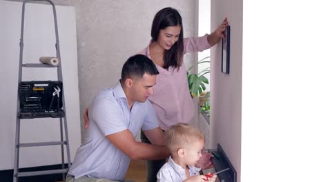 dad-teaches-son-to-bolt-shelf-to-wall-with-screwdriver-and-mother-near-hanging-picture-during-renovation-apartment