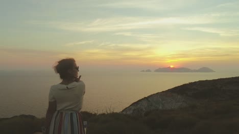 Hermosa-mujer-joven-con-sombrero-de-moda-colorido-vestido-con-la-falda-y-flores-mirando-la-puesta-de-sol-en-montaña-de-la-isla-de-Ponza-Italia.-Tiro-de-Drone-aéreo.