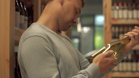 Attractive-guy-carefully-examines-the-composition-on-the-label-of-a-white-wine-bottle-in-liquor-store.-Wine-bottles-are-standing-on-wooden-shelves-in-alcohol-shop.