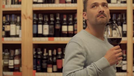 Handsome-man-standing-with-glass-of-red-wine-in-liquor-store.-Woman-in-the-background-is-choosing-a-wine-bottle