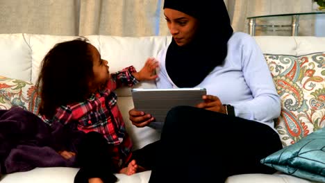 Madre-joven-usando-una-tableta-digital-con-su-hija-en-el-sofá-en-casa-4k