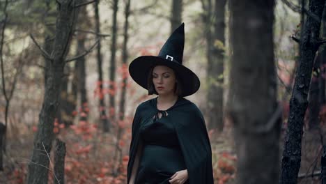 Woman-as-black-witch-walks-between-trees-in-autumn-forest.Girl-in-long-dress,-cape,-fairy-hat.-Halloween-concept,-cosplay-dressing-up