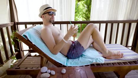 joven-lleva-shorts-de-verano,-sombrero-de-paja-y-gafas-de-sol,-relajarse-en-una-tumbona-en-una-tienda-de-campaña-y-beber-jugo-de