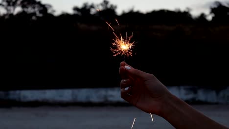 Fading-sparkler-in-a-female-hand-with-white-manicure-in-the-evening-on-the-beach,-close-up,-slow-motion