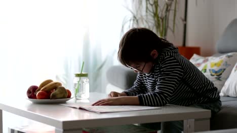 Preescolar-niño-con-gafas,-escribir-tareas-en-casa-en-el-soleado-salón