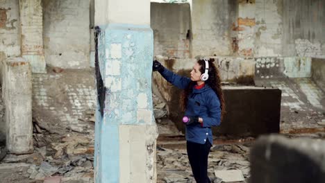 Tilt-Up-Shot-schlankes-Girl-moderne-Maler-Zeichnung-Graffiti-auf-hohen-Säule-in-zerstörten-Lager-mit-Aerosol-Farbe-und-Musikhören-mit-Kopfhörer.