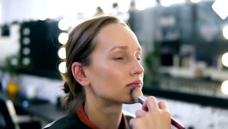 Hand-of-make-up-artist-applying-tone-on-a-model's-face-using-a-big-brush.-Beautiful-caucasian-blonde-woman.-Blurred-make-up-studio-on-the-background