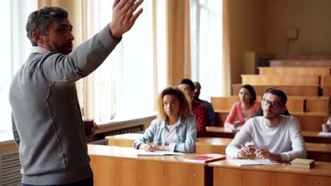 Bärtiger-Mann-Professor-liest-Vortrag-sprechen-und-Gestikulieren,-während-Studenten-hören-und-schreiben-sitzen-an-Tischen-in-geräumigen-Universität-Klassenzimmer-sind.