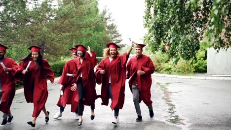 Excited-graduating-students-are-running-with-diplomas-on-campus-territory-wearing-gowns-and-traditional-hats,-it-is-raining.-Higher-education-and-happiness-concept.