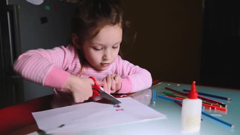 Cute-little-preschool-European-girl-child-in-pink-sweater-cutting-her-drawing-out-of-paper-sheet,-talking-to-someone