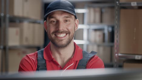 Portrait-of-Uniformed-Worker-Using-Personal-Computer-while-Sitting-at-His-Desk-in-the-Warehouse.-In-the-Background,-Room-with-Shelves-Full-of-Cardboard-Box-Packages-Ready-For-Shipping.