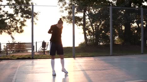 Caucasian-sporty-young-woman-in-hoodie-and-shorts-warming-up-in-morning-on-basketball-court.-Sun-shines-on-the-background