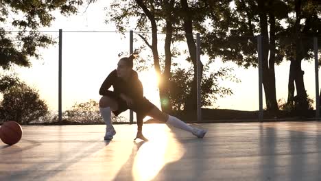 Caucasian-young-sporty-woman-in-hoodie-and-shorts-warming-up-in-morning-on-basketball-court.-Stretching-her-legs-before-basketball-training.-Sun-shines-on-the-background