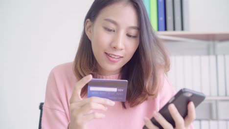 Beautiful-Asian-woman-using-smartphone-buying-online-shopping-by-credit-card-while-wear-sweater-sitting-on-desk-in-living-room-at-home.-Lifestyle-woman-at-home-concept.