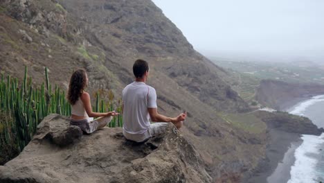 Un-hombre-y-una-mujer-sentada-en-la-cima-de-una-montaña-mirando-el-mar-sentado-en-una-piedra-meditando-en-posición-de-loto.-La-vista-desde-la-parte-posterior.-Islas-Canarias