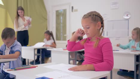 Retrato-de-colegiala-en-la-elaboración-de-lecciones-en-aula-luz-sentado-detrás-de-escritorio-fondo-de-compañeros-de-clase-y-joven-maestro