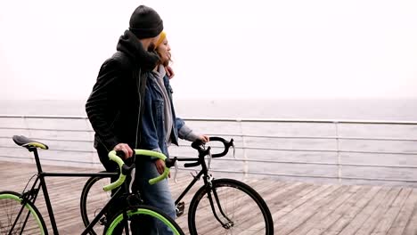 Hermosa-pareja-sonriente-de-los-hipsters-jóvenes-caminando-juntos-abrazando-con-sus-bicicletas-cerca-del-mar-en-día-de-otoño.-Chica-joven-de-sombrero-amarillo-en-algo.-Caminando-por-cubierta-de-madera-en-día-nublado.-Cierre-para-arriba