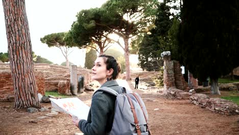 Back-view-of-beautiful-tourist-girl-with-backpack-and-map-exploring-ancient-sunny-amphitheater-ruins-in-Ostia,-Italy.