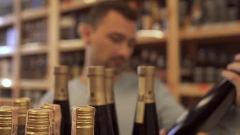 Customer-reading-the-composition-of-the-wine-on-the-bottle-label,-nods-approvingly-and-going-away-taking-the-wine-bottle-with-him.-Guy-who-choosing-the-wine-in-the-store-is-out-of-focus.