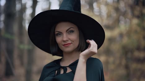 Portrait-of-young-pretty-witch-in-cap-on-autumn-forest-background.-Halloween-concept,-cosplay-dressing-up.-Slow-motion