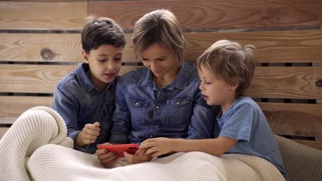 Joven-mujer-caucásica-con-dos-hijos-se-divierten-usando-el-teléfono-inteligente-relajarse-en-el-sofá,-los-niños-felices-disfrutan-de-pasar-tiempo-en-casa-con-mamá.-Videojuegos