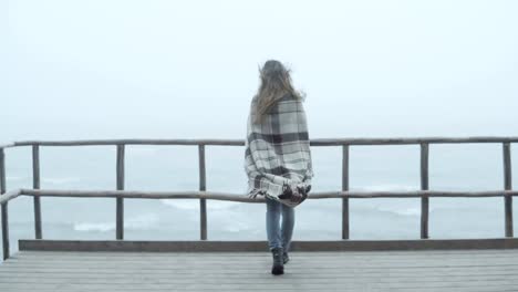 Back-view-of-young-woman-with-plaid-standing-on-the-wooden-pier-and-looking-afar.-Female-spends-time-alone-in-foggy-day