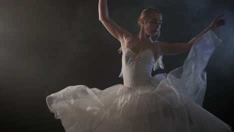 Tiro-medio-de-una-hermosa-bailarina-joven-girando-graciosamente-en-el-centro-de-atención.-Ella-vestida-de-vestido-blanco-luminoso-moderno-tutu.-Mujer-sensual-en-el-escenario-negro-humo.-Cámara-lenta