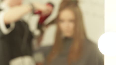 Blurred-reflection-in-mirror-hairdresser-making-woman-hairstyling-long-hair-with-dryer-and-brush-in-beauty-salon.-Hairstyling-fashion-model-in-dressing-room