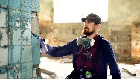 Serious-bearded-guy-is-concentrated-on-painting-graffiti-on-old-dirty-column-inside-abandoned-building-with-aerosol-paint.-Man-is-wearing-casual-clothes-and-gloves.