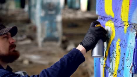 Graffiti-painter-is-squatting-near-column-in-old-empty-building-and-painting-graffiti-with-spray-paint-creating-beautiful-image.-Creative-people-and-modern-art-concept.