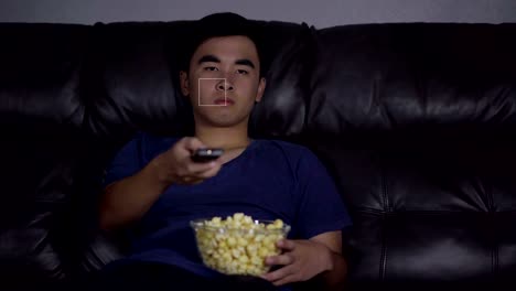 young-man-holding-remote-control-and-watching-TV-while-sitting-on-sofa-at-night