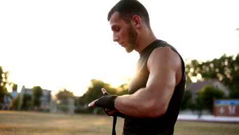 Side-view-of-a-young,-bearded-boxer-preparing-for-training-outdoors,-wrapping-black-bandage-while-stading-on-the-stadium