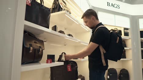 Young-male-customer-is-examining-bags-in-a-shop,-goods-are-on-racks
