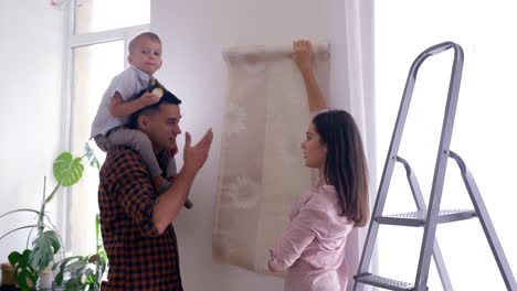 happy-family-doing-repairs-in-new-home,-joyful-Mom-and-daddy-with-son-on-shoulders-chose-wallpaper-in-apartment