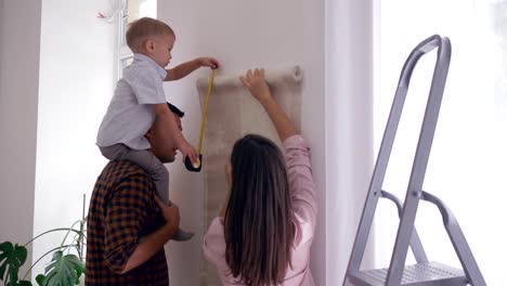 family-renovation-in-room,-young-mother-and-father-with-boy-on-shoulders-glue-new-wallpaper-on-wall