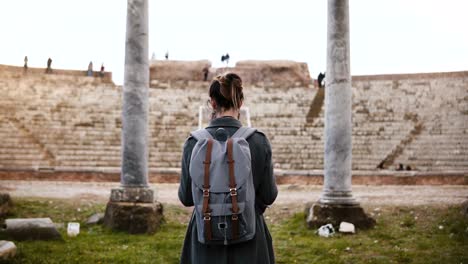 Back-view-of-beautiful-young-female-tourist-with-backpack-and-map-exploring-ancient-amphitheater-ruins-in-Ostia,-Italy.