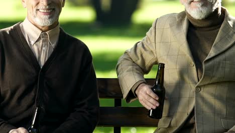 Smiling-aged-men-showing-beer-bottles,-sitting-on-bench,-non-alcoholic-beverage