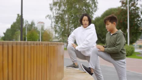 Asiática-Madre-e-Hijo-Estirando-Piernas-durante-el-Entrenamiento-al-Aire-Libre