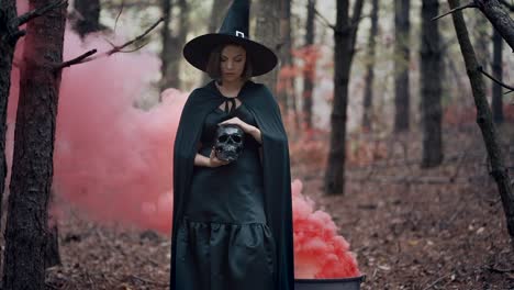 Black-witch-holding-skull-in-hands,-stroking-it-on-autumn-smoke-forest-background.-Horror,-halloween,-cosplay-holiday,-magic-concept