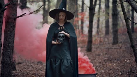 Schwarze-Hexe-hält-Schädel-in-den-Händen,-streichelt-es-auf-Herbst-Rauch-Wald-Hintergrund.-Horror,-Halloween,-Cosplay-Urlaub,-magisches-Konzept