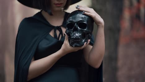 Black-witch-holding-skull-in-hands,-stroking-it-on-autumn-forest-background.-Horror,-halloween,-cosplay-holiday,-magic-concept