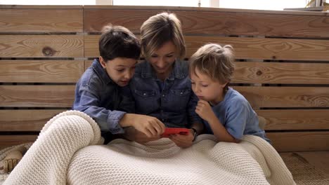 Joven-mujer-caucásica-con-dos-hijos-se-divierten-usando-el-teléfono-inteligente-relajarse-en-el-sofá,-los-niños-felices-disfrutan-de-pasar-tiempo-en-casa-con-mamá.-Videojuegos