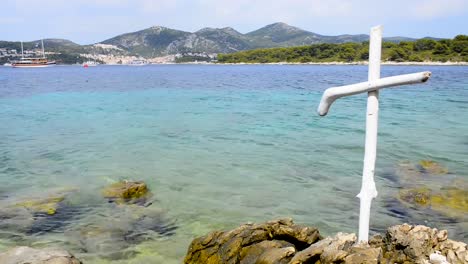 Christlichen-weißen-Holzkreuz-am-felsigen-Ufer-des-blauen-Meeres.-Küste-der-Insel-Hvar-und-Hafen-in-der-Altstadt-von-Hvar.